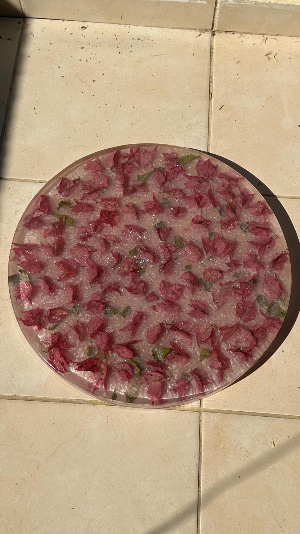 The round resin top on its own, seen from above on a tiled floor: dried pink bougainvillea petals and small green leaves in pearly pink resin